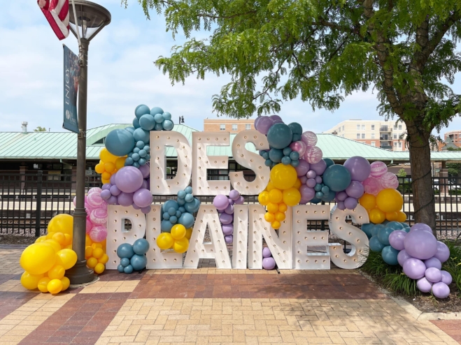 Shows a large sculpture that says "Des plaines" in theatre letters with balloons all around